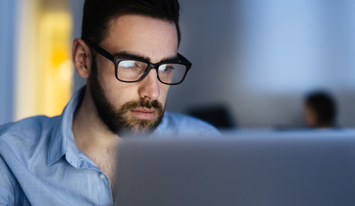 young male with beard professional in an office wearing glasses working and focusing on laptop